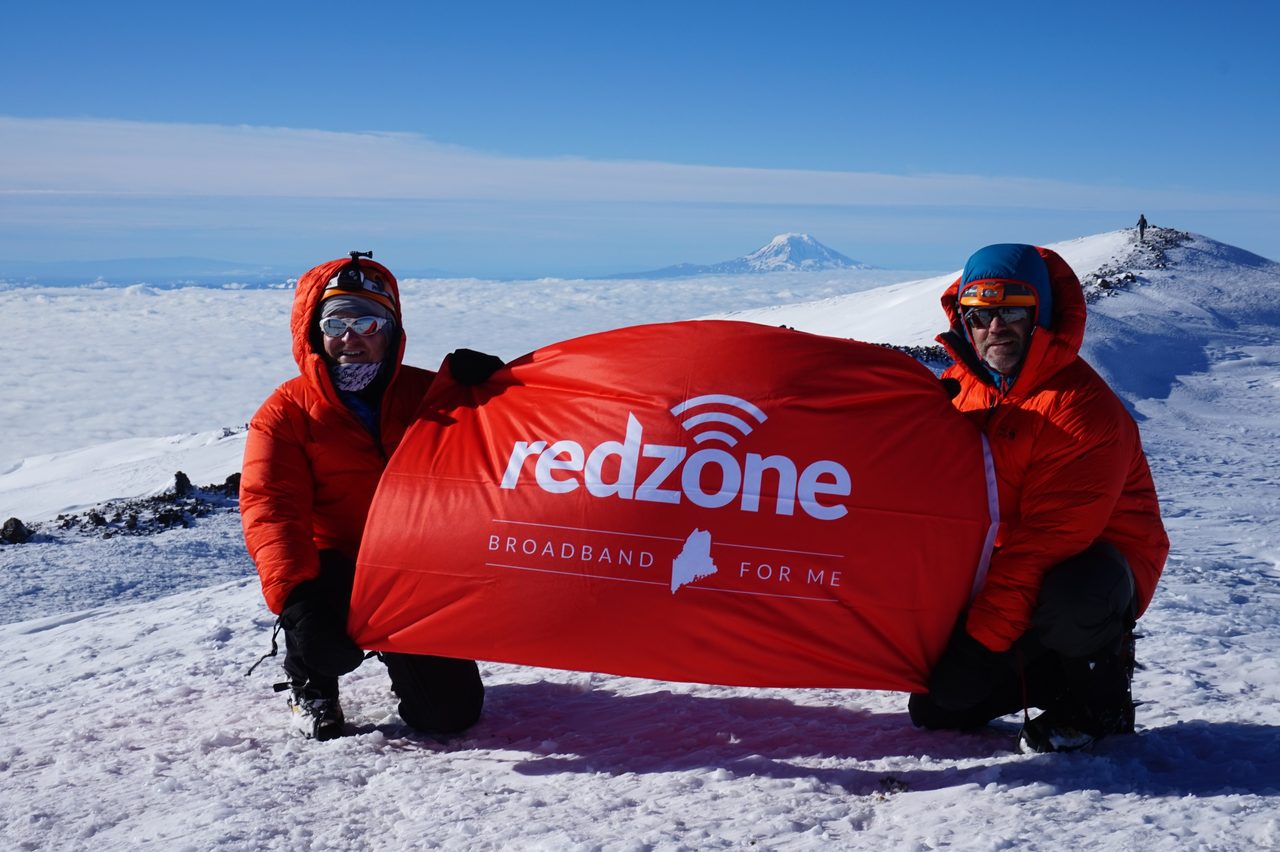 Two climbers in red jackets holding a Redzone banner on the snowy summit of Mt. Rainier