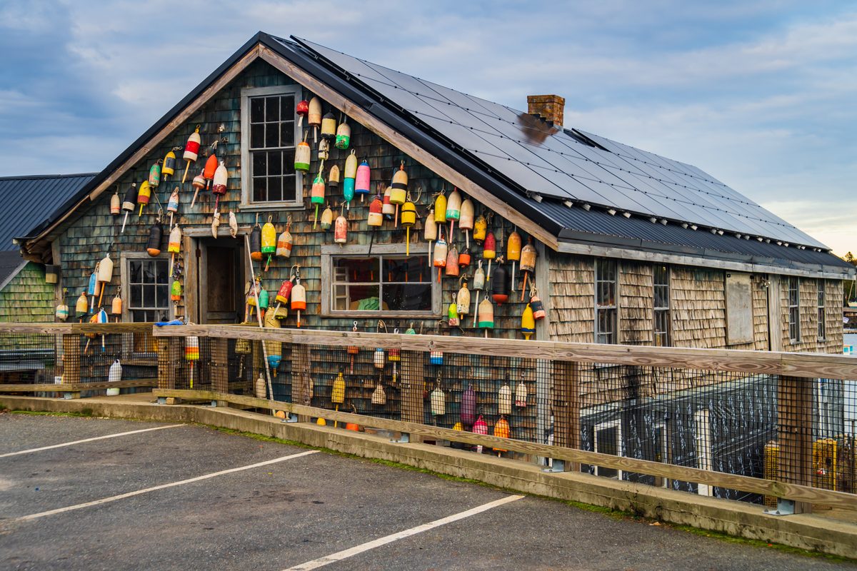 A shingled lobster building on a Maine wharf, covered in colorful buoys, with solar panels on the roof